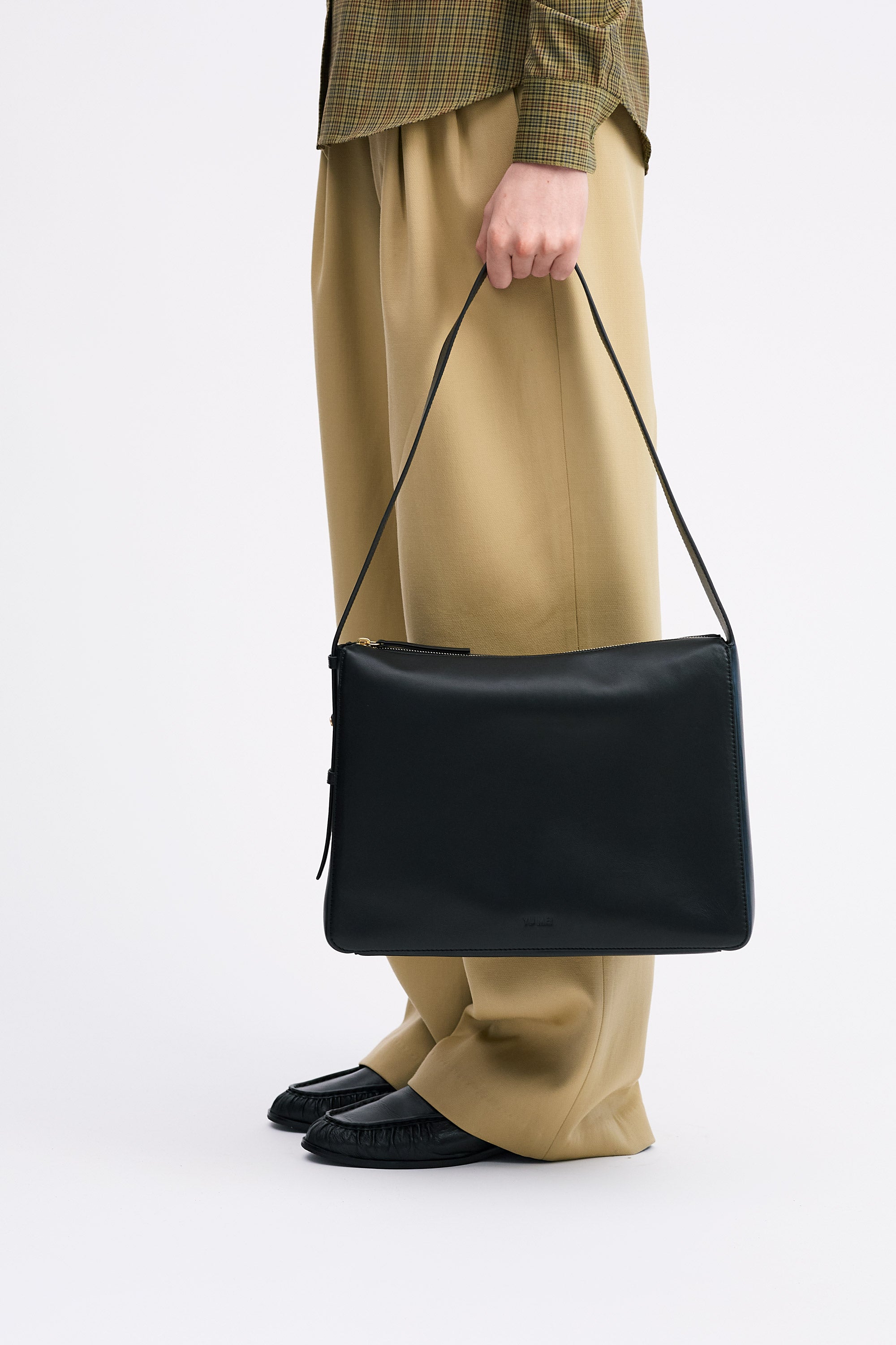 Model holding the Charlie Bag in Black lambskin in hand, wearing a check shirt and tan pants